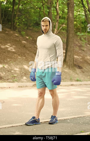 Grand jour arrive. Athlète homme concentré sur le visage avec des gants de boxe sport pratique prêt punch fond nature. Boxer hooded motivé pour réussir. Boxer sportif formation avec des gants de boxe. Banque D'Images