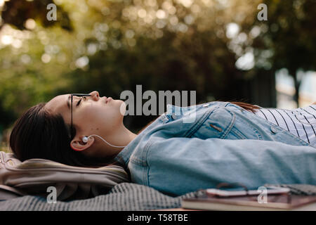 Gros plan d'une femme dormant dans un parc à l'écoute de musique. Niveau du sol tourné d'une jeune femme étendue sur le sol portant des lunettes avec des livres à côté d'elle Banque D'Images