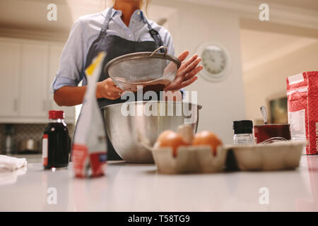 Chef sasser la farine dans un bol pour faire un gâteau au chocolat. Femme portant un tablier la cuisson dans la cuisine avec des ingrédients autour. Banque D'Images