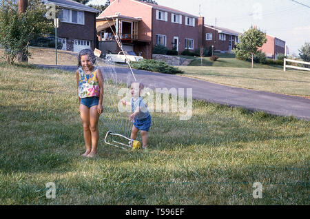 Années 1970, historiques, les enfants s'amuser en plein air...une jeune fille ayant une douche au moyen d'un sprinkleur le jardin, regardait avec amusement par son petit frère pour tester l'eau, USA. Banque D'Images