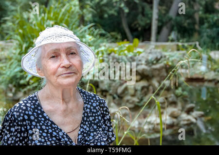 Closeup portrait of a senior woman sereine dans un tricot chapeau blanc, se tenant debout à l'extérieur en été Banque D'Images