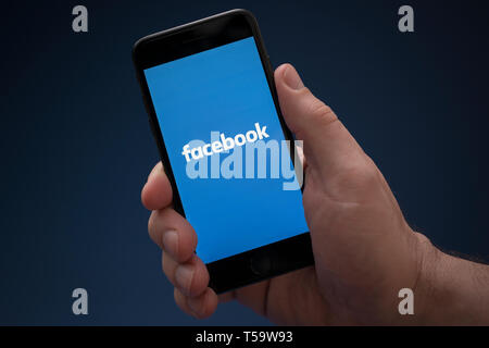 Un homme se penche sur son iPhone qui affiche le logo de Facebook (usage éditorial uniquement). Banque D'Images