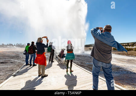 Les touristes de prendre une photo de geyser de ruche qu'il éclate contre un ciel bleu clair le jour d'ouverture de Parc National de Yellowstone comme le dégel du printemps s'ouvre le parc pour les visiteurs après un long hiver, le 19 avril 2019 dans la région de Yellowstone, Wyoming. Banque D'Images