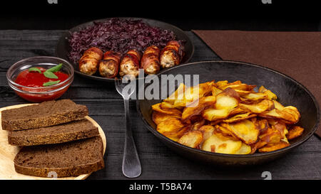 Pommes de terre rôties dans le pot, des morceaux de pain noir de seigle sur une assiette de service en bois, des saucisses au bacon et compotée de chou sur une zone rustique noir Banque D'Images