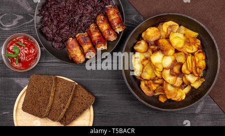 Pommes de terre rôties dans le pot, des morceaux de pain noir de seigle sur une assiette de service en bois, des saucisses au bacon et compotée de chou sur une zone rustique noir Banque D'Images