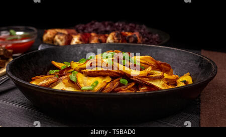 Croustilles de pommes de terre frites avec des greens dans un pot, en face de des saucisses et compotée de chou, on a black background rustique. Banque D'Images