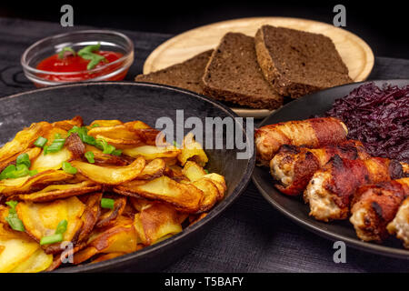 Pommes de terre rôties dans le pot, des morceaux de pain noir de seigle sur une assiette de service en bois, des saucisses au bacon et compotée de chou sur une zone rustique noir Banque D'Images