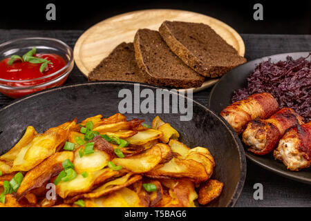 Pommes de terre rôties dans le pot, des morceaux de pain noir de seigle sur une assiette de service en bois, des saucisses au bacon et compotée de chou sur une zone rustique noir Banque D'Images