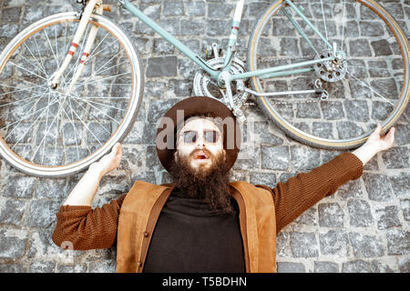 Portrait d'un homme barbu élégant dans hat lying with retro vélo sur la chaussée, la rue vue du dessus Banque D'Images