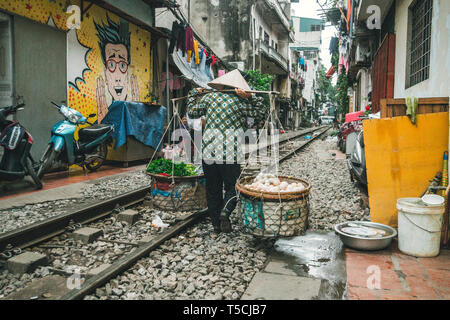 Hanoi. Le Vietnam. 13 Janvier 2019 : femme porte une chape traditionnelle avec de la nourriture par chemin de fer le long de rue étroite avec des maisons Banque D'Images