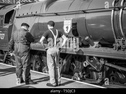 Vue arrière en noir et blanc de l'équipe de locomotive à vapeur se tenant sur la plate-forme de chemin de fer sous le soleil inspectant le côté du moteur d'époque du Royaume-Uni. Banque D'Images