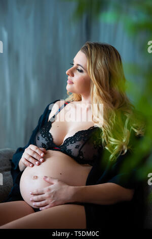 En photoshoot enceintes style loft. Belle mère enceinte avec des cheveux blonds est assis sur un canapé et rêveur regarde par la fenêtre et sur le ventre. Banque D'Images