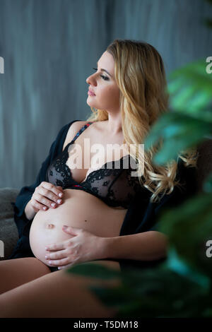 En photoshoot enceintes style loft. Belle mère enceinte avec des cheveux blonds est assis sur un canapé et rêveur regarde par la fenêtre et sur le ventre. Banque D'Images