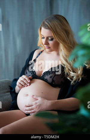 En photoshoot enceintes style loft. Belle mère enceinte avec des cheveux blonds est assis sur un canapé et rêveur regarde par la fenêtre et sur le ventre. Banque D'Images