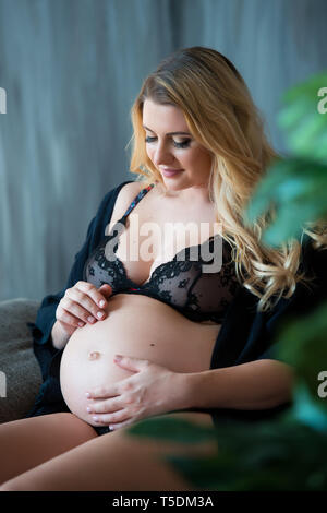 En photoshoot enceintes style loft. Belle mère enceinte avec des cheveux blonds est assis sur un canapé et rêveur regarde par la fenêtre et sur le ventre. Banque D'Images