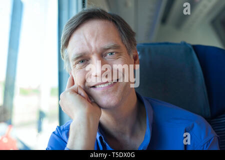 Handsome Young man in blue shirt assis sur le train de banlieue en plein jour, smiling at camera Banque D'Images