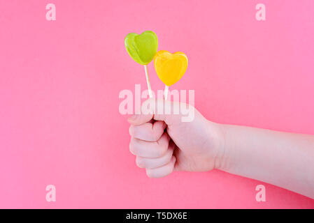 En forme de coeur jaune et vert lollipops. Dans la main de l'enfant. Sur fond rose Banque D'Images