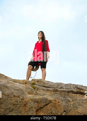 Image ado asiatique caucasienne Biracial girl with long hair en chemise rouge debout sur le sommet rocheux élevé souriant sur camera Banque D'Images