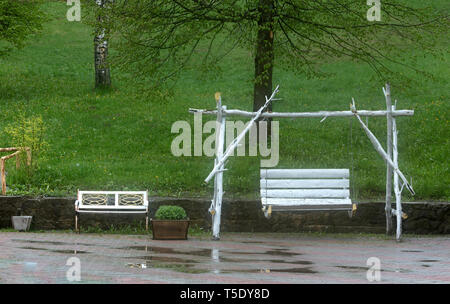 Balançoires blanc dans le jardin après la pluie d'été sont enveloppées dans des vapeurs de brouillard dans le contexte de l'herbe verte juteuse et de flaques sur le trottoir t Banque D'Images