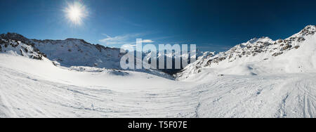 Vue panoramique vers le bas dans la vallée enneigée de montagnes alpines sur fond de ciel bleu avec soleil Banque D'Images