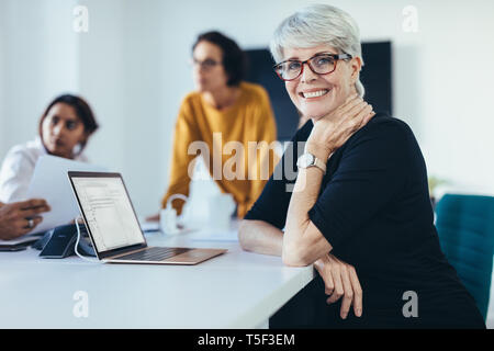 Succès Mid adult woman sitting lors d'une réunion avec des collègues dans la salle. Avec des collègues de sexe féminin dans la salle de conférence à la caméra et au sourire. Banque D'Images