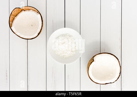 Deux moitiés de noix de coco et flocons de noix de coco dans un bol en porcelaine blanche sur le panneau blanc arrière-plan avec copie espace. Vue d'en haut. Banque D'Images