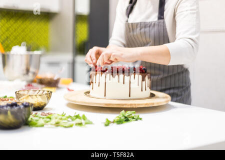 Pâtissier décore avec des baies un biscuit gâteau au chocolat blanc et crème. Cake se dresse sur un support en bois sur un tableau blanc. Le concept de Banque D'Images