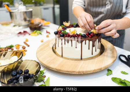 Pâtissier décore avec des baies un biscuit gâteau au chocolat blanc et crème. Cake se dresse sur un support en bois sur un tableau blanc. Le concept de Banque D'Images