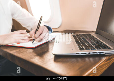 Close-up of a woman's hand writing dans un bloc-notes sur l'arrière-plan d'un ordinateur portable et de l'intérieur d'un avion Banque D'Images