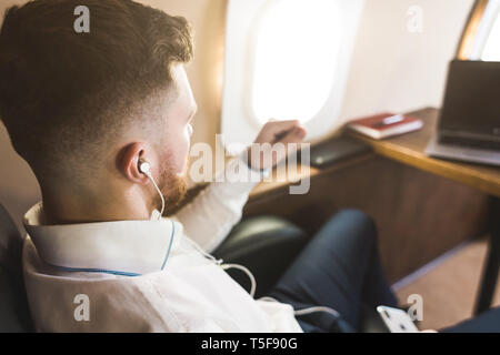 Jeune homme d'affaires prospère et attrayante est au repos et à l'écoute de musique fonctionne alors qu'il était assis dans le fauteuil de son avion d'affaires privé Banque D'Images