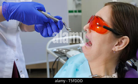 Portrait de jeune femme peur d'enlever une dent assis dans fauteuil dentaire pendant que le médecin se tient à côté du patient, tenant dans les mains. forceps Banque D'Images