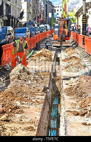 Embouteillage et entrepreneur de travaux routiers à creuser une tranchée dans les routes et rues London West End pour les câbles à l'aide de mini digger derrière des barrières London England UK Banque D'Images