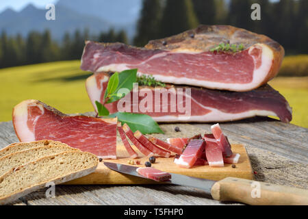 Bacon Tyrolien du Sud typique snack avec pain de seigle local allongé sur une table rustique en face des alpages et montagnes des Alpes Banque D'Images