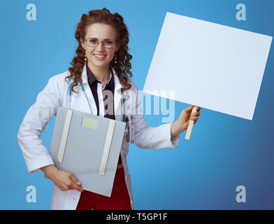 Femme médecin élégant souriant en bue chemise, pantalon rouge et blanc robe médical avec l'échelle montrant le poids de l'étiquette vierge isolé sur fond bleu. Banque D'Images