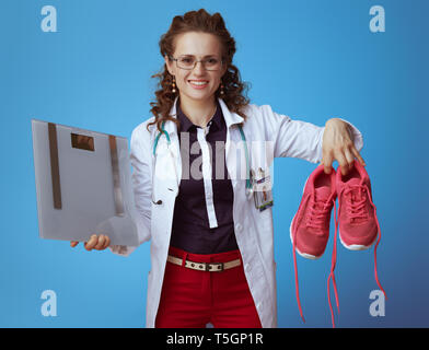 Femme médecin suitcase en bue chemise, pantalon rouge et blanc robe médical avec l'échelle montrant le poids des chaussures de conditionnement physique isolé sur fond bleu. Banque D'Images