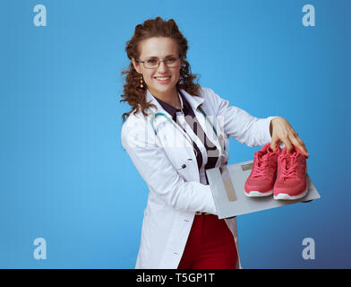 Femme médecin élégant souriant en bue chemise, pantalon rouge et blanc robe médical montrant l'échelle de poids et de forme physique sneakers isolé sur bleu. Banque D'Images