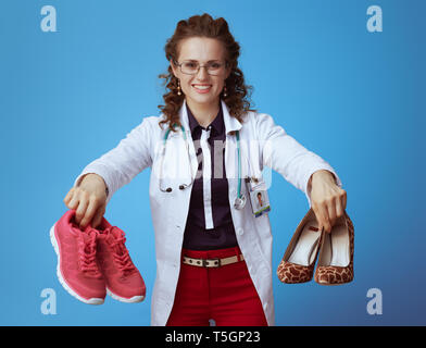 Médecin moderne smiling woman in bue chemise, pantalon rouge et blanc robe médical fitness montrant les espadrilles et hautes chaussures de talon contre bac bleu Banque D'Images