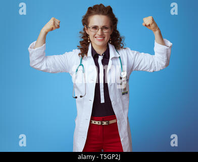 Femme médecin moderne souriant en bue chemise, pantalon rouge et blanc robe médical montrant biceps isolé sur bleu. Banque D'Images