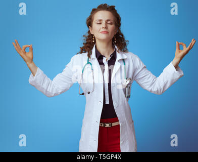 Portrait de femme médecin moderne et relaxant en bue chemise, pantalon rouge et blanc robe médical faisant du yoga sur fond bleu. Banque D'Images