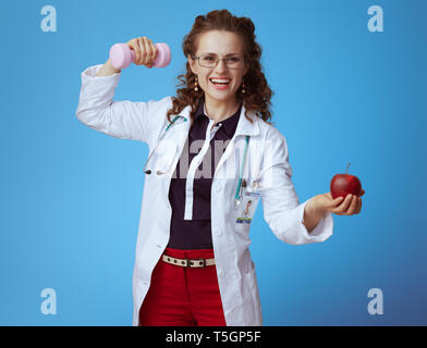 Femme médecin moderne heureux en bue chemise, pantalon rouge et blanc robe médical avec haltère et apple montrant biceps isolé sur bleu. Banque D'Images