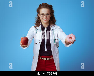 Femme médecin élégant souriant en bue chemise, pantalon rouge et blanc robe médical montrant apple et donut isolé sur fond bleu. Banque D'Images