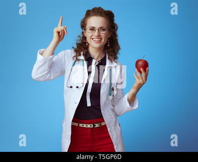 Femme médecin moderne souriant en bue chemise, pantalon rouge et blanc robe médical montrant une pomme s'idée isolé sur bleu. Banque D'Images