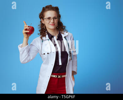 Femme médecin suitcase en bue chemise, pantalon rouge et blanc robe médical avec une pomme et se lève le doigt sur fond bleu. Banque D'Images