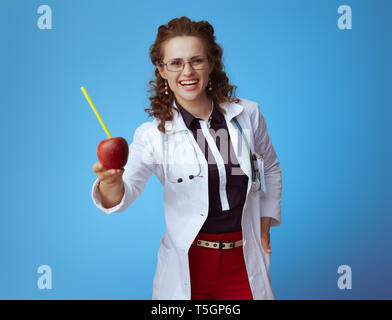 Femme médecin moderne souriant en bue chemise, pantalon rouge et blanc robe médical donnant une pomme verte avec tube à cocktails sur fond bleu. Banque D'Images