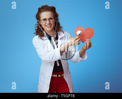 Femme médecin moderne souriant en bue chemise, pantalon rouge et blanc robe médical stéthoscope écoute avec papier rouge coeur isolé sur fond bleu. Banque D'Images