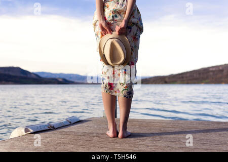 Vue arrière de barefoot young woman standing on jetty holding summer hat, vue partielle Banque D'Images