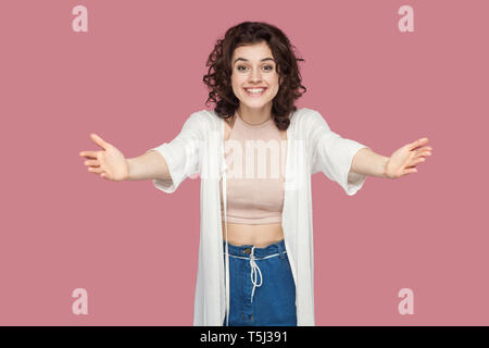 Portrait of happy brunette jeune femme avec des poils frisés dans style casual debout avec les bras levés, à hug, sourire à pleines dents et à la recherche à Banque D'Images