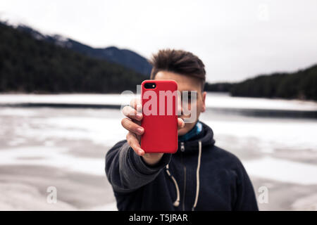 Jeune homme tenant téléphone cellulaire rouge à un lac en hiver Banque D'Images