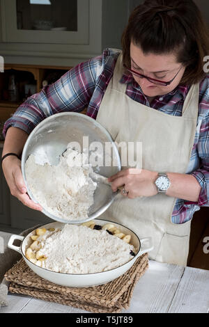 Femme portant tablier, debout dans une cuisine fraîche, verser le mélange de crumble sur un moule à tarte rempli de fruits. Banque D'Images