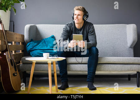 Smiling Young man with guitar, tablette et écouteurs sitting on couch Banque D'Images